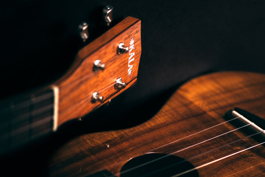 Closeup of Kala ukulele headstock and body including tuners, nut, fretboard, strings, bridge, saddle, and sound hole.